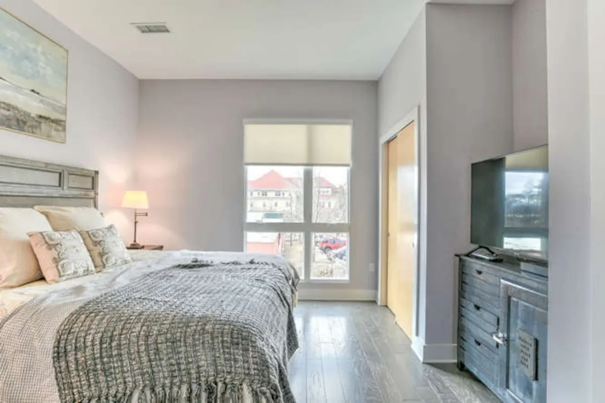 This image shows a serene bedroom setting with a cozy bed adorned with decorative pillows, a large window that allows natural light to flood the space, a nightstand with a lamp, and a TV unit. The view through the window subtly hints at a connection to the outdoors, suggesting the presence of the balcony beyond.