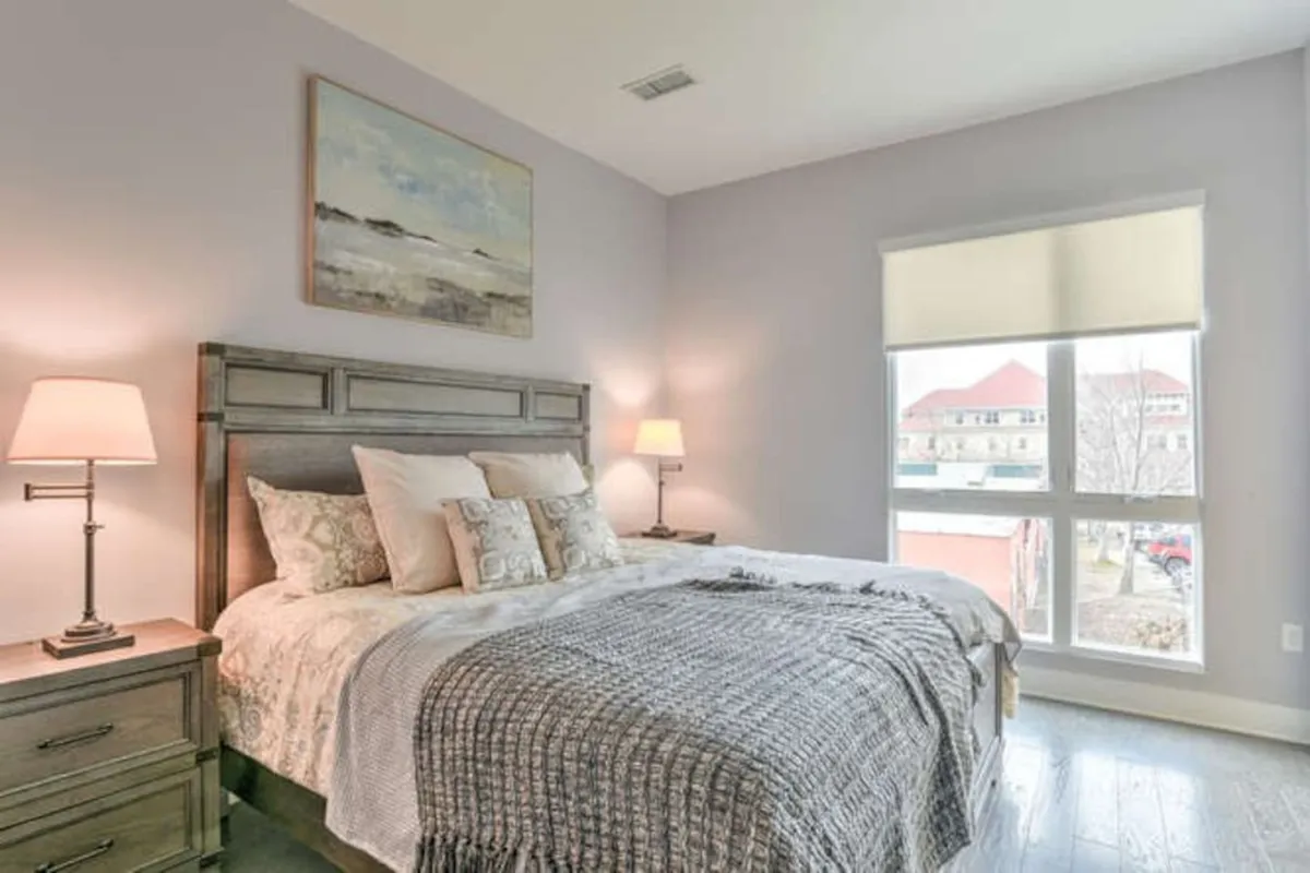 The image features a tranquil bedroom with a comfortable bed, soothing artwork, nightstands with lamps, and large windows that brighten the room. The windows are a central element, reinforcing the connection to outdoor spaces like the balcony.