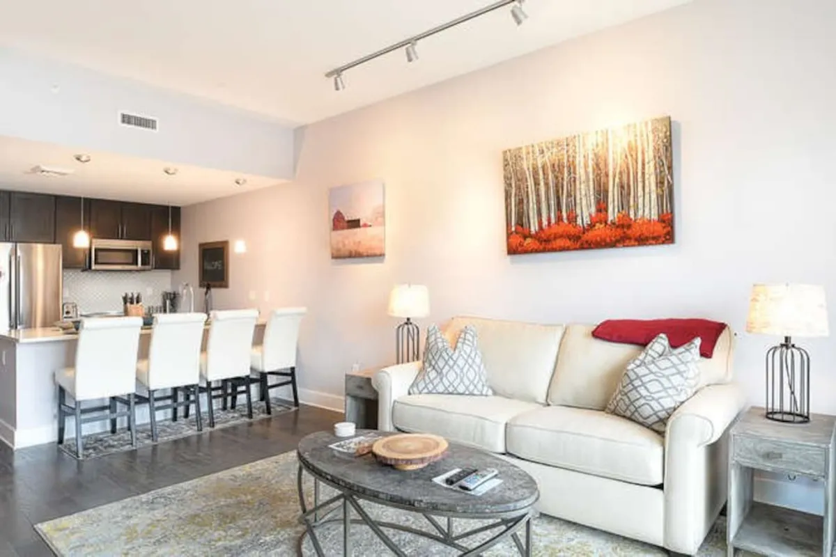 A welcoming open-concept living area featuring a plush sofa, vibrant artwork, and a modern kitchen island with bar stools. The space is filled with warm light and designed for both relaxation and social gatherings, with a seamless connection between the living, dining, and kitchen zones.