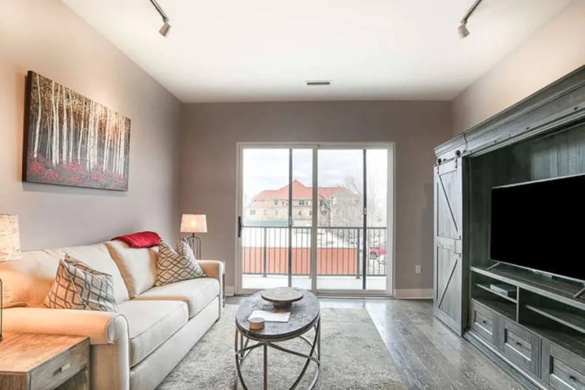 A comfortable living room arrangement with a large, inviting sofa, eye-catching artwork on the wall, and expansive windows that flood the space with natural light. A chic coffee table anchors the room, offering a spot for drinks, games, or conversation.