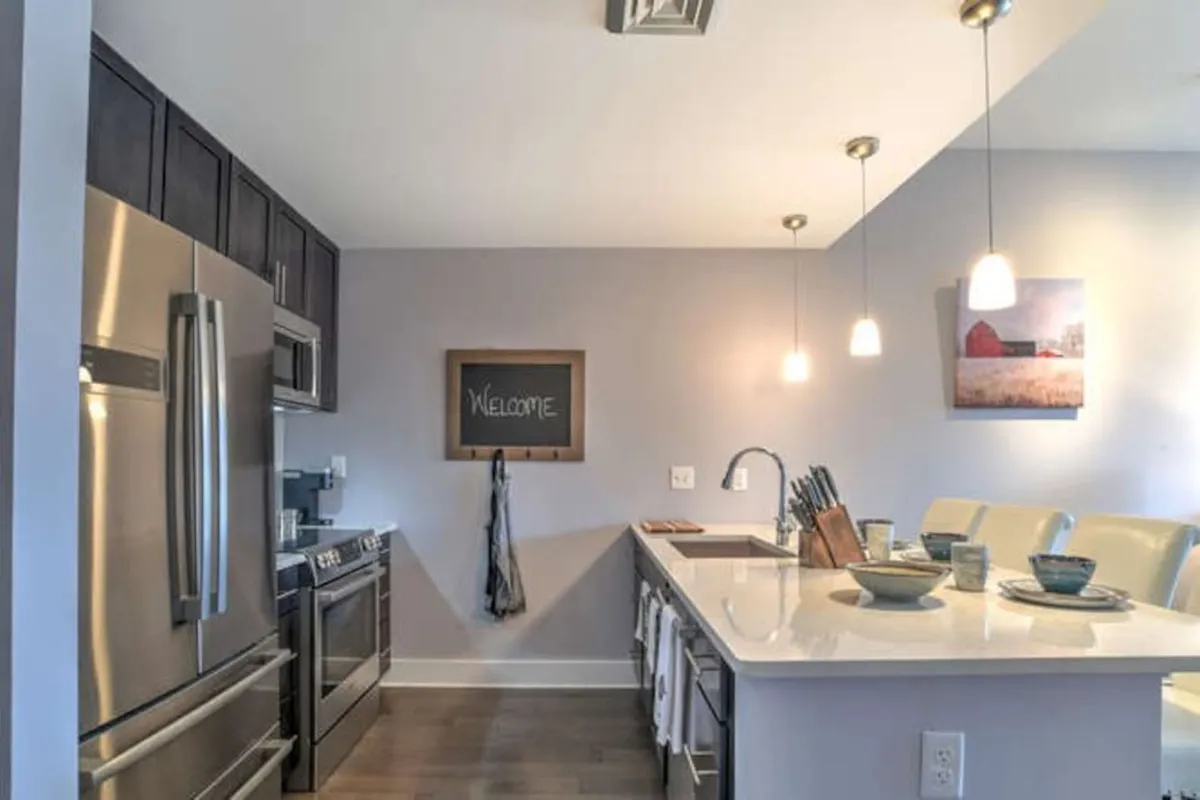 A bright, open kitchen space featuring sleek stainless steel appliances, a crisp white countertop, and a friendly chalkboard sign welcoming guests. The adjoining dining area is set for a meal with bowls ready, evoking a sense of hospitality and community gathering.
