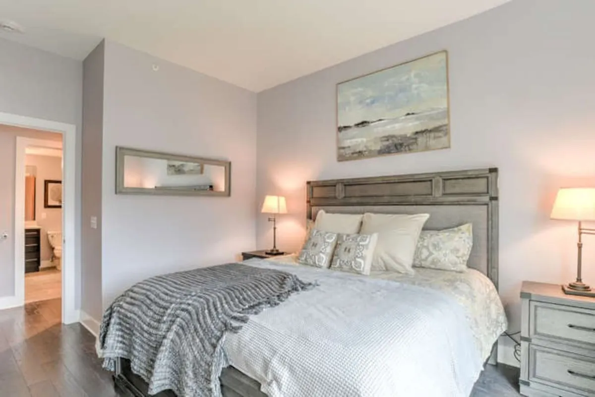 This image features a king-sized bed adorned with decorative pillows, flanked by nightstands with illuminated lamps and complemented by tasteful artwork above the headboard. The soft natural light filtering through the room conveys a tranquil and restful ambiance.